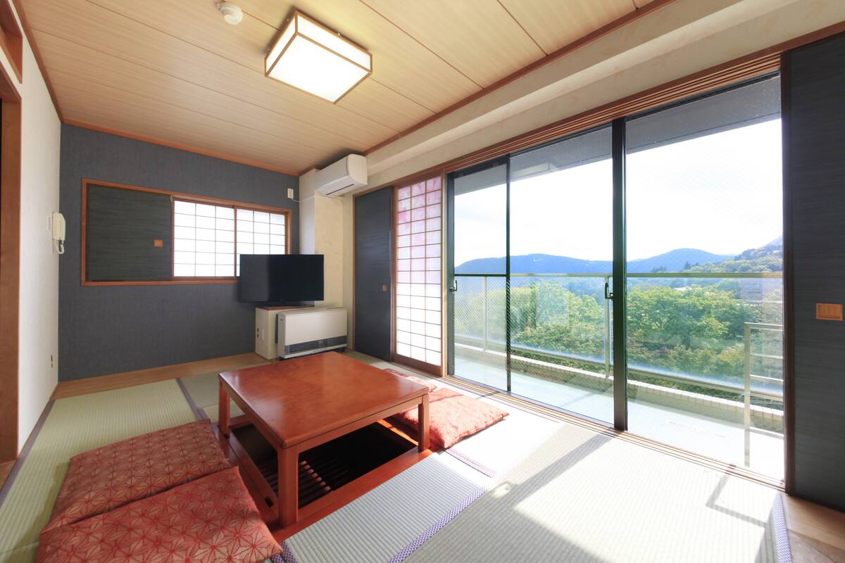A traditional Japanese room with tatami flooring, a sliding door to a balcony, a television, and a low wooden table.