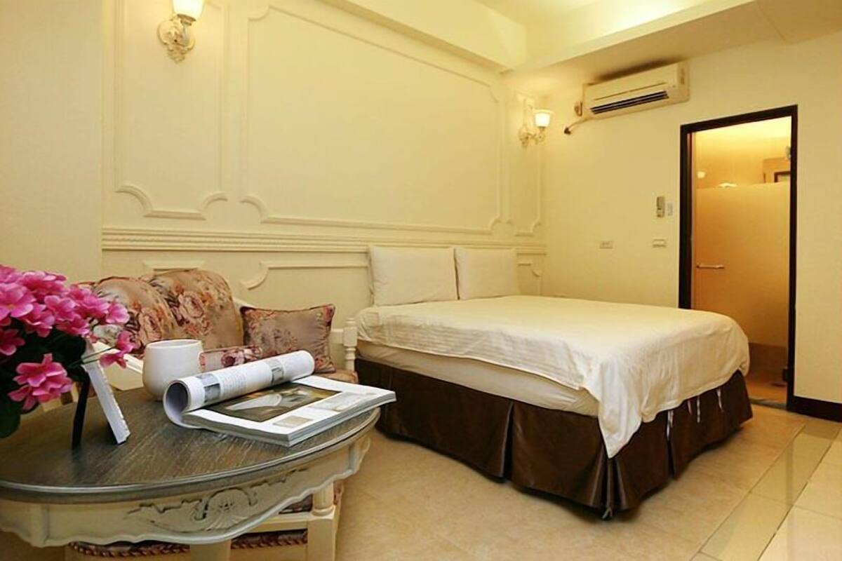 A hotel room with a neatly made bed, a small table with a vase of flowers, and a door leading to another room.