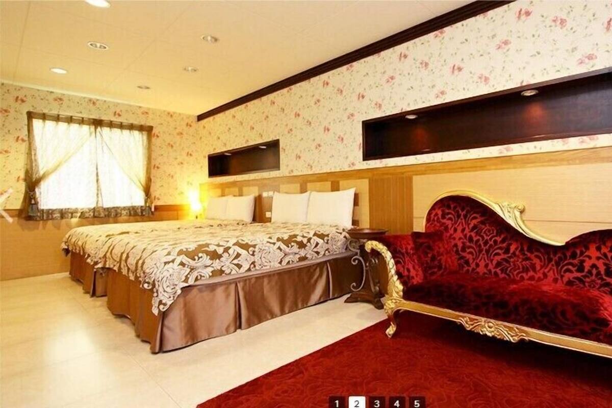 A hotel room with a large bed, a red velvet sofa, floral wallpaper, and a flat-screen TV mounted on the wall.