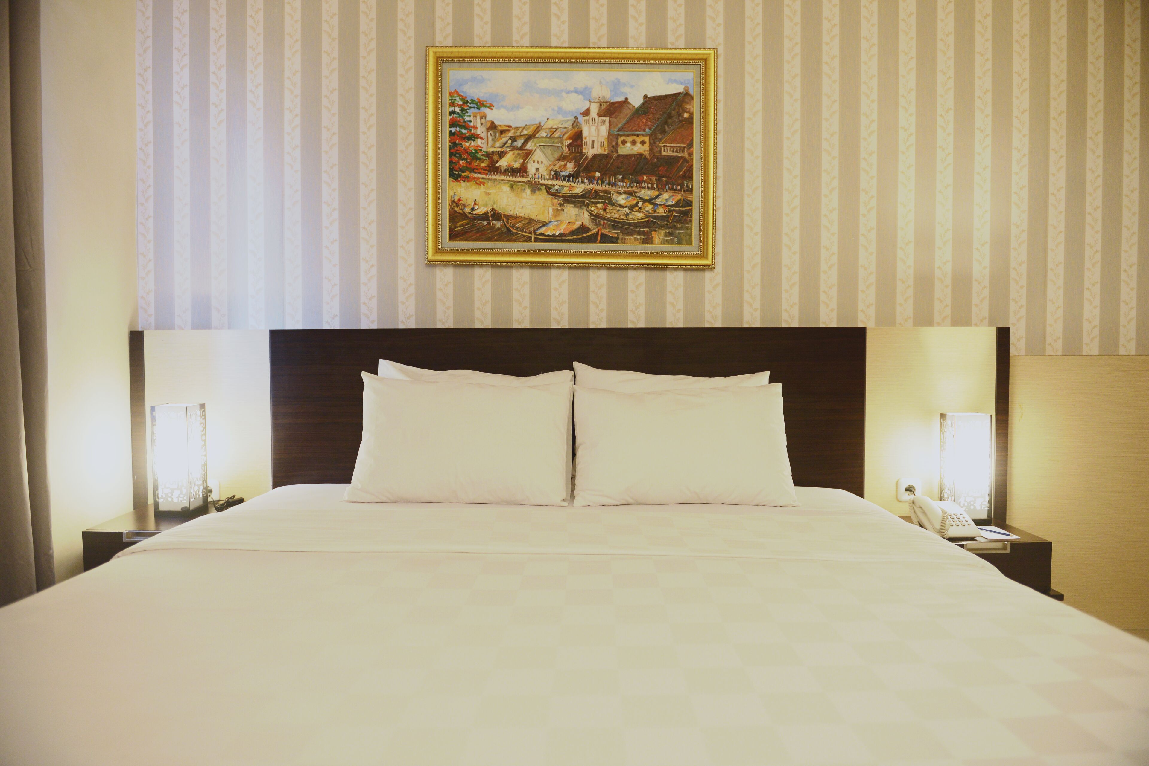 A hotel room with a large bed, two bedside tables with lamps, a framed painting on the wall, and striped wallpaper.