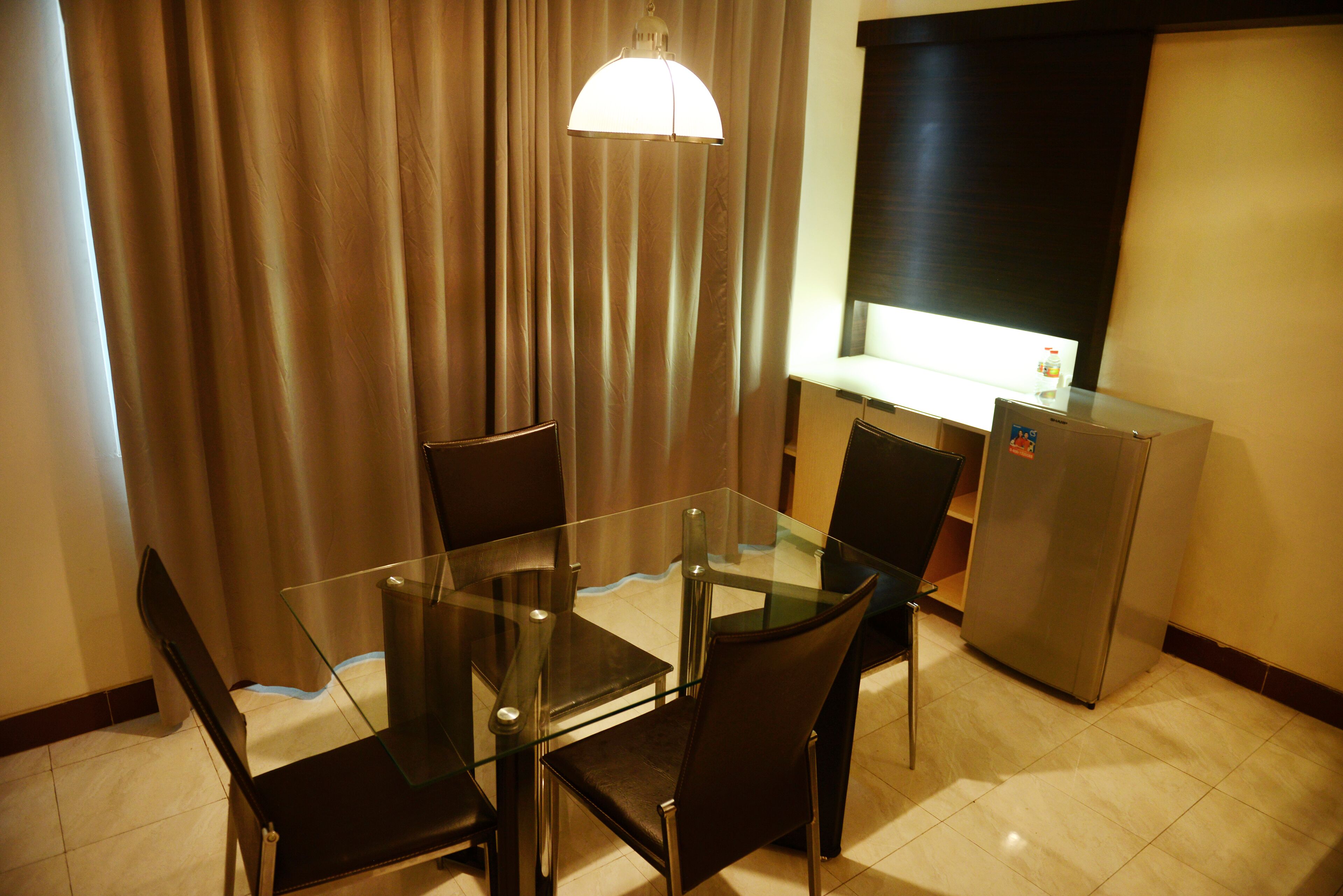 A hotel room with a glass dining table, four chairs, a refrigerator, and a curtain.