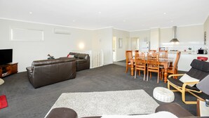 A living room with a brown sofa, a dining table with chairs, and a kitchen area in the background. - Bayside (Aldinga Beach)