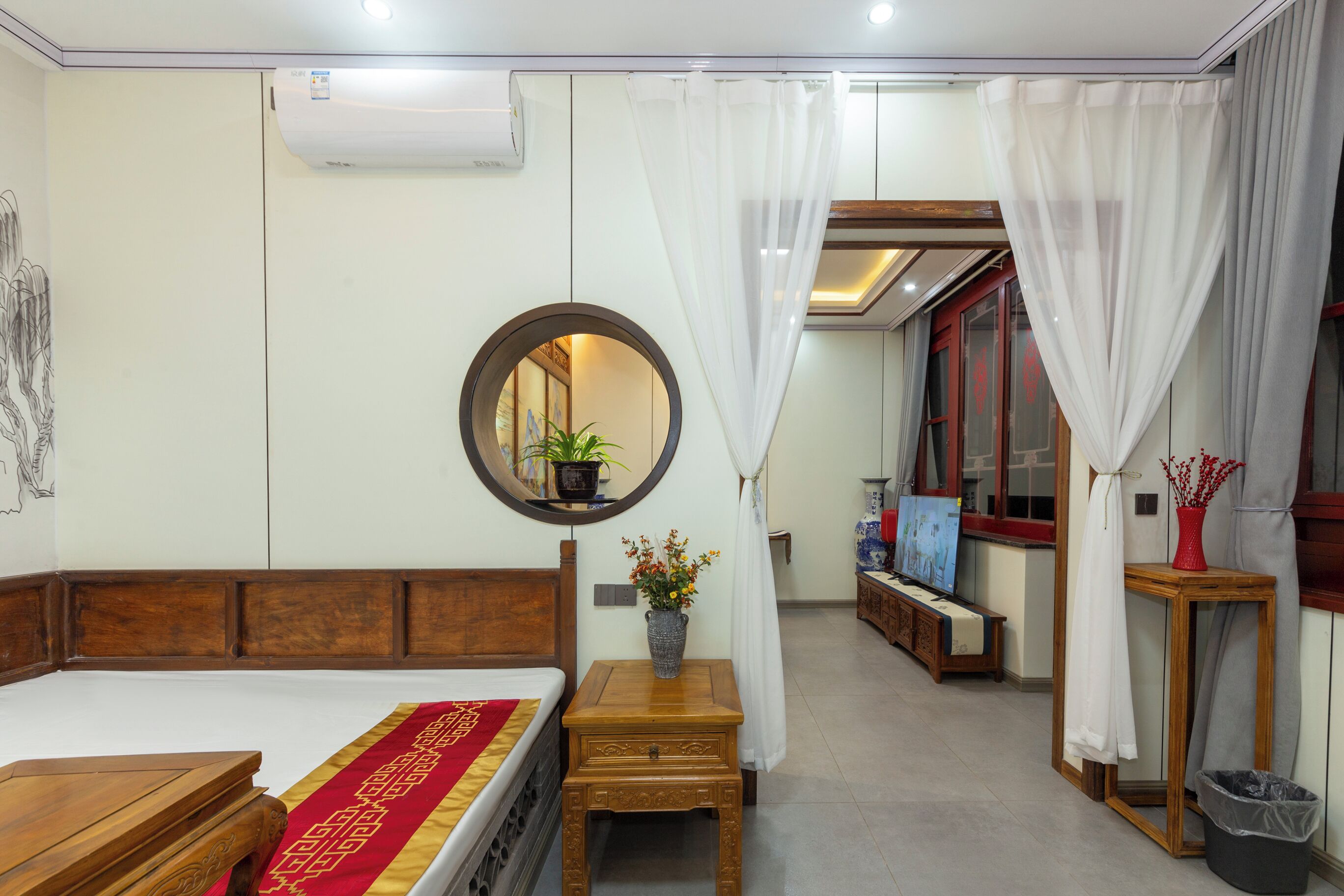 A bedroom with a wooden bed, a bedside table with a vase, a mirror, and a window with curtains.