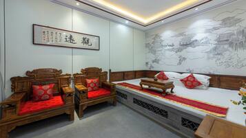 Habitación tradicional con una cama de madera, dos sillas de madera, un cojín rojo y una pared decorada con una obra de caligrafía enmarcada.
