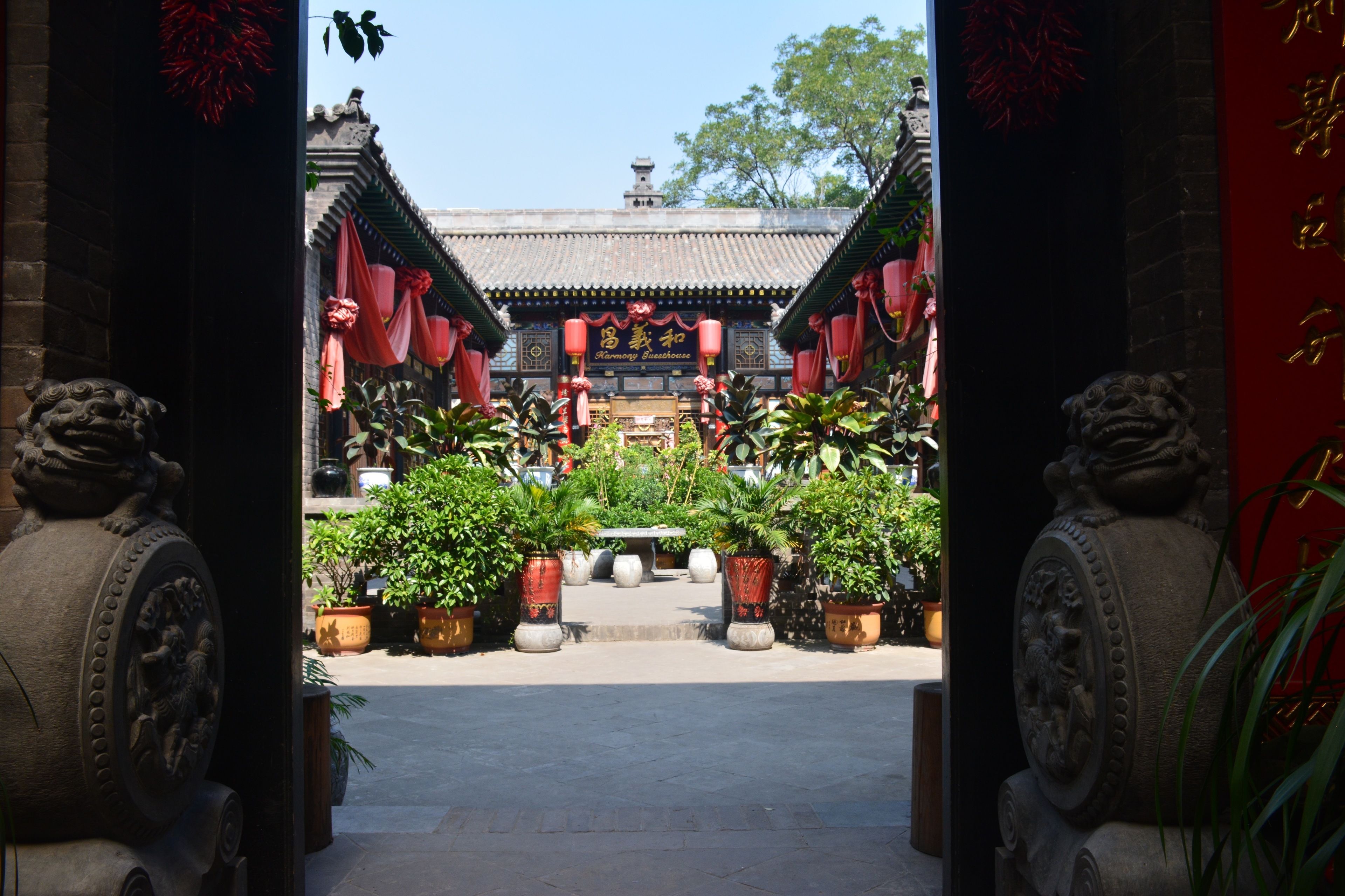 A traditional Chinese courtyard with ornate lion statues at the entrance, potted plants, and a tiled roof.