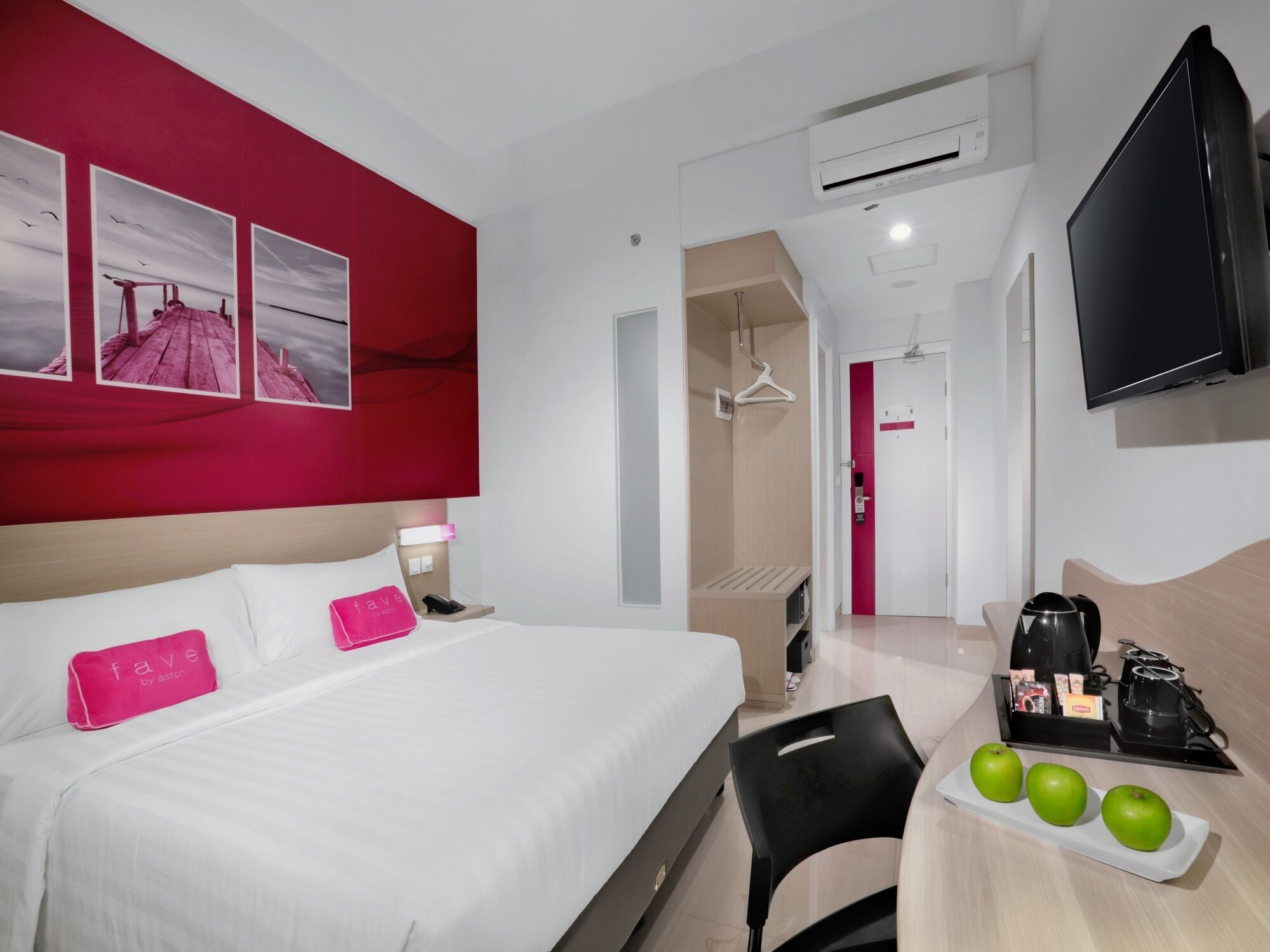 Habitación de hotel con una cama grande, un escritorio con una bandeja de manzanas verdes, una silla negra, un televisor y una pared de acento roja con cuadros.
