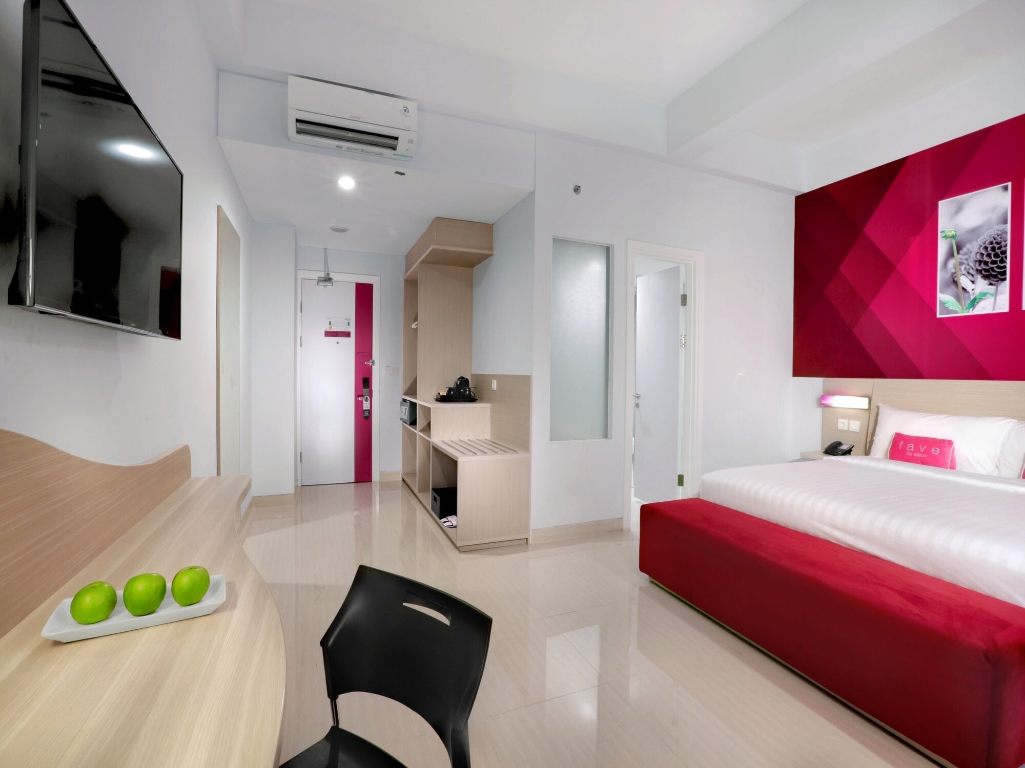 A modern hotel room with a bed, a desk with apples, a chair, a TV, and a red accent wall.