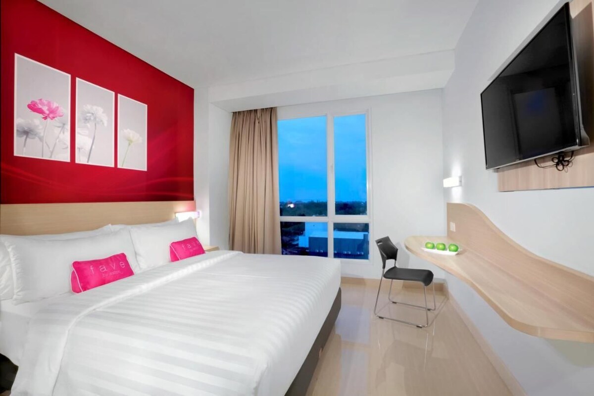 A hotel room with a large bed, a TV mounted on the wall, a window with a view, and a desk with a chair.