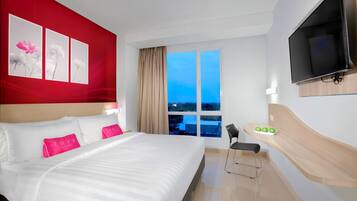 Ett hotellrum med en stor säng, en tv som är monterad på väggen, ett skrivbord med en stol och ett fönster med utsikt över staden i skymningen.