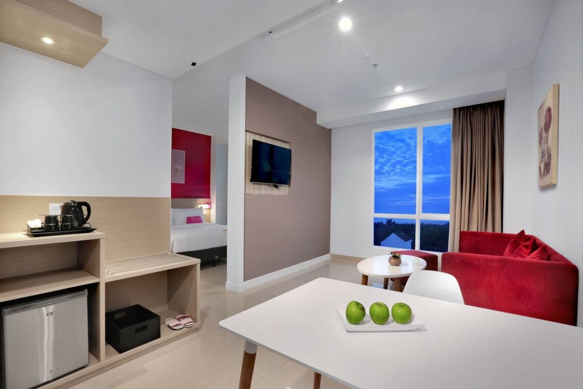A modern hotel room with a kitchenette, a dining area with a table and chairs, a red sofa, and a large window with a view of the sky.