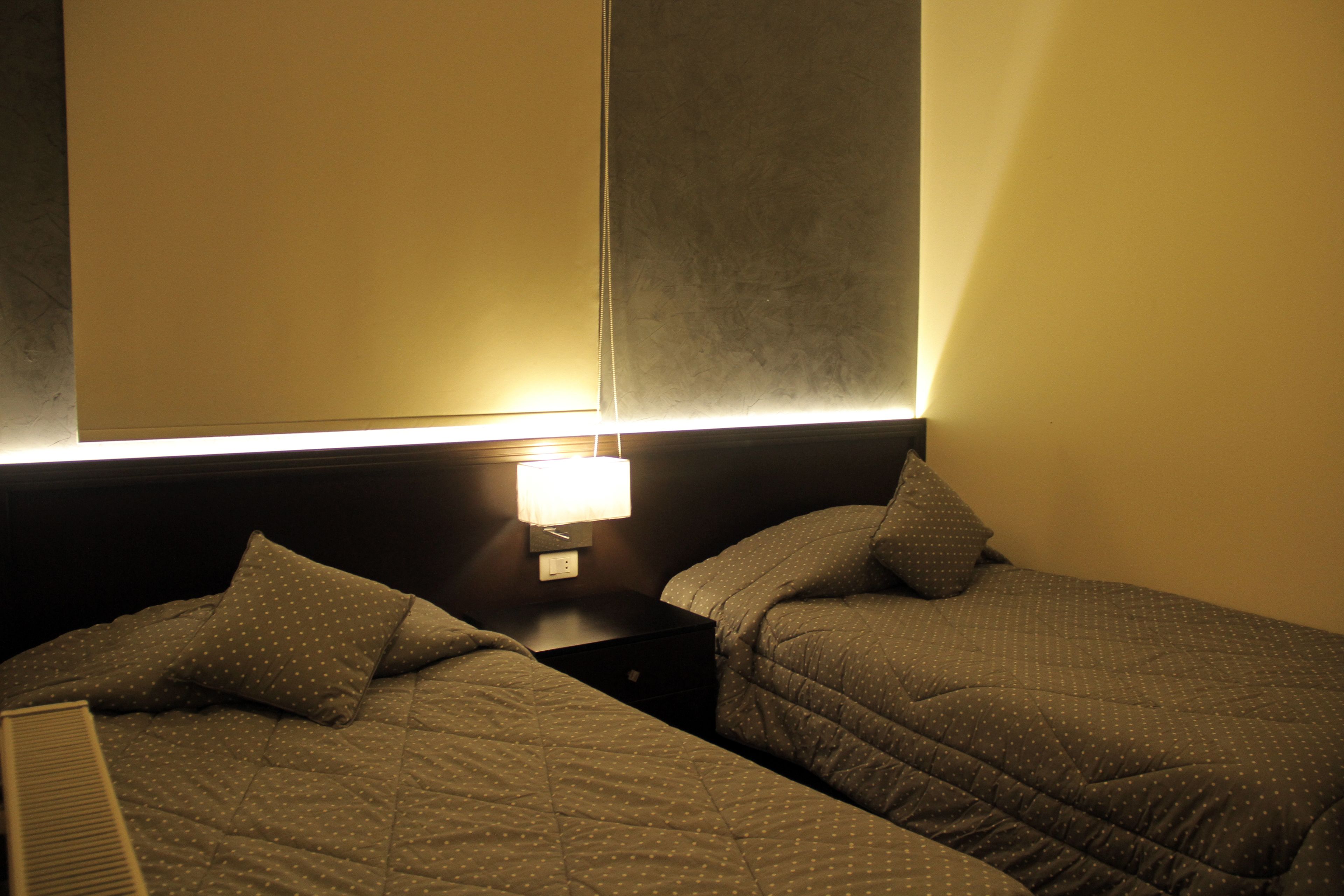 A hotel room with two single beds, each with a pillow and a quilted blanket. There is a nightstand with a lamp between the beds.