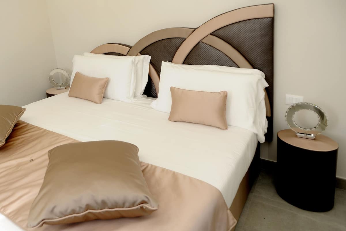 A neatly made bed with beige bedding and pillows, a round mirror on a side table, and a headboard with a woven design.