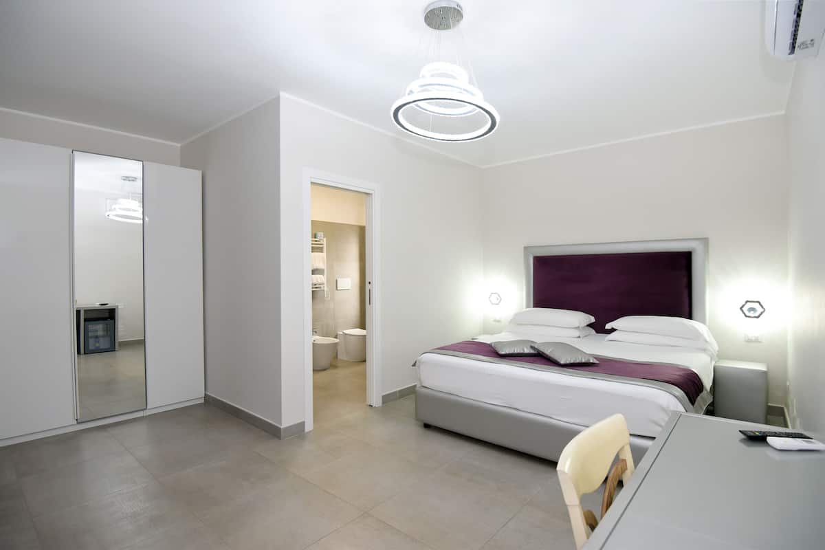 A modern hotel room with a bed, bedside tables, a desk, and a chair. There is a bathroom visible through an open door.
