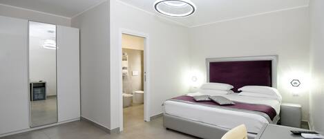Une chambre d’hôtel moderne, équipée d’un lit, de tables de chevet, d’un bureau et d’une chaise. On peut apercevoir la salle de bain à travers une porte ouverte.