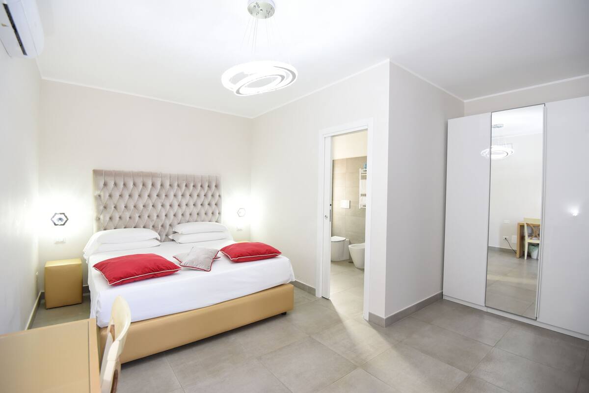 A modern bedroom with a tufted headboard, a bed with white bedding and red pillows, a bedside table, and a wardrobe with a mirror.