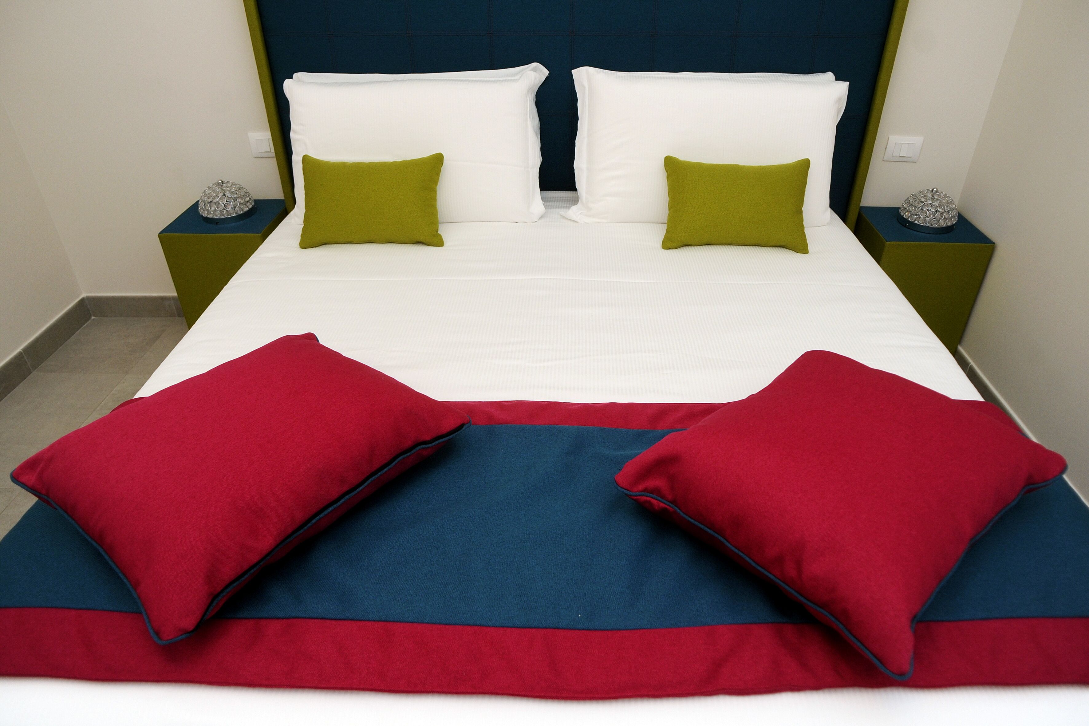 A neatly made bed with white and green pillows, a red and blue blanket, and two decorative items on the nightstands.