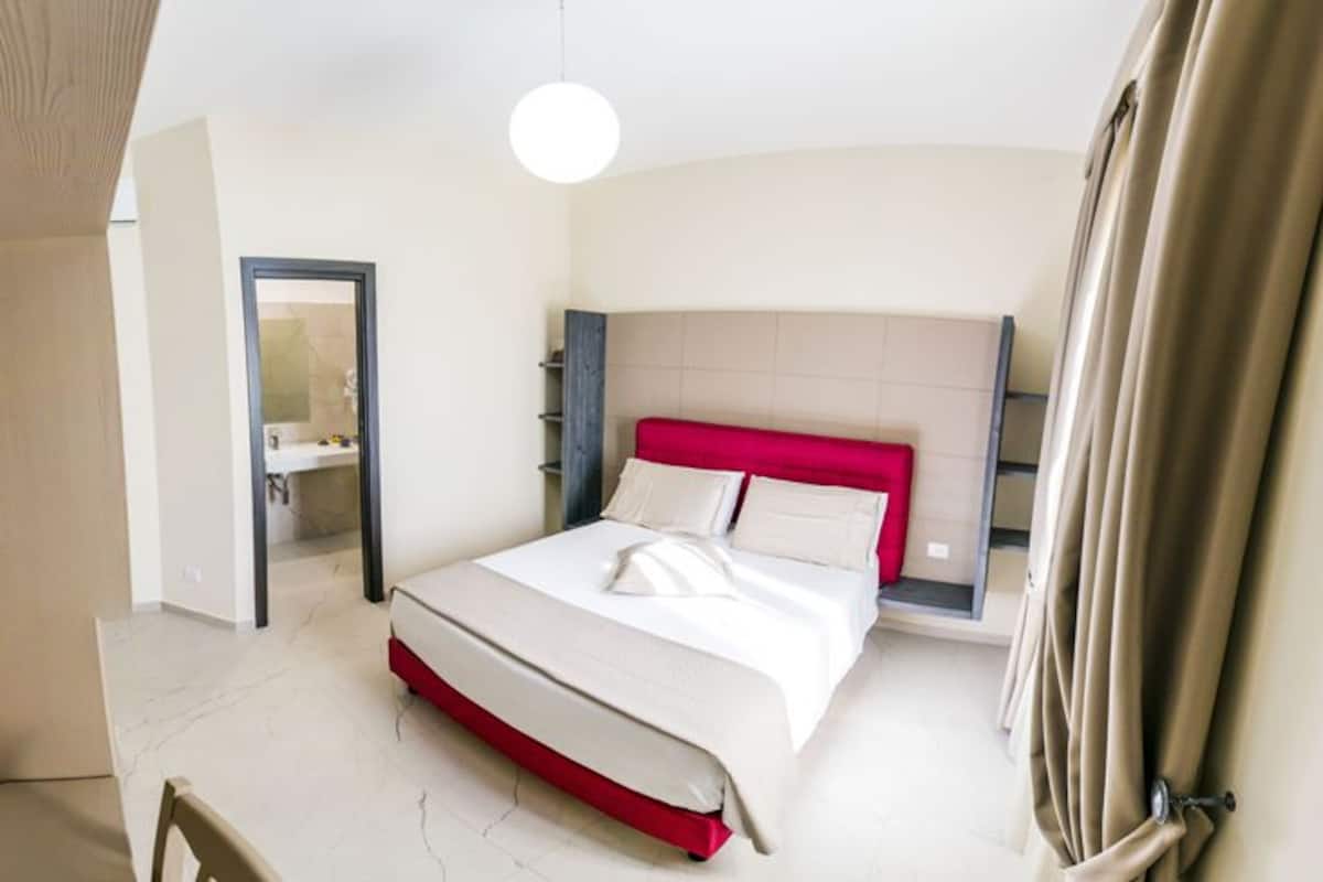 A modern hotel room with a large bed, a bedside table, a mirror, and a door leading to a bathroom.