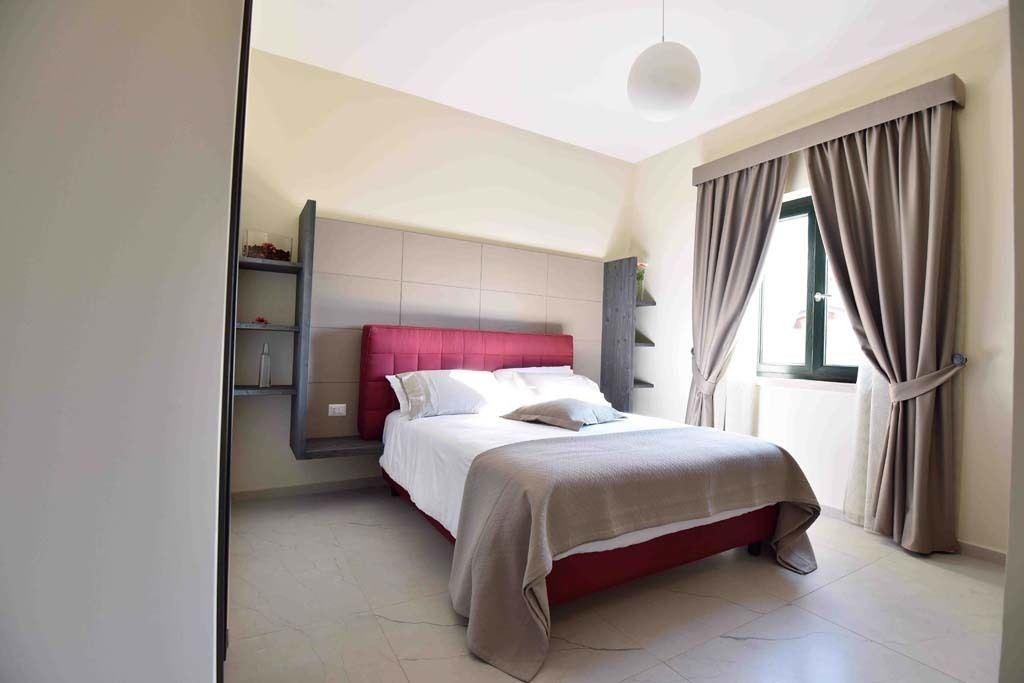 A modern bedroom with a red headboard, a large bed, a bedside table, and a window with curtains.