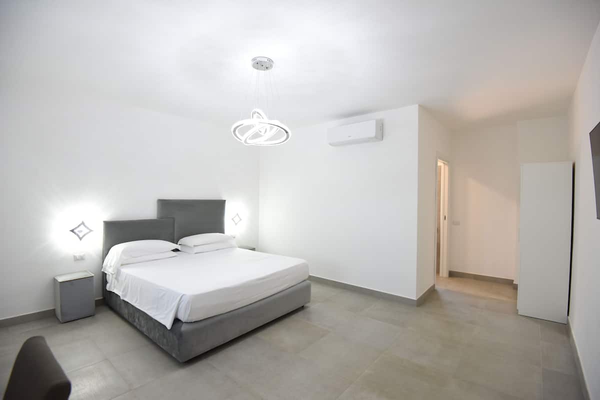 A modern bedroom with a bed, bedside tables, a hanging light fixture, and a door leading to another room.