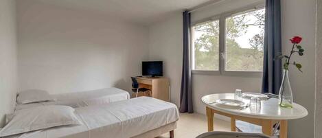Ein Schlafzimmer mit Bett, Schreibtisch mit Fernseher, Esstisch mit Stühlen und einem Fenster mit Vorhängen.