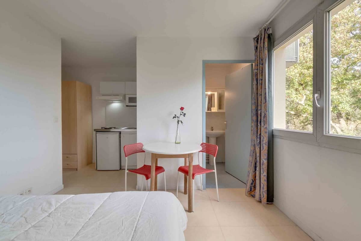 Ein Schlafzimmer mit einem Bett, einem kleinen Esstisch mit Stühlen, einer Küchenzeile im Hintergrund und einem Fenster mit Vorhängen.