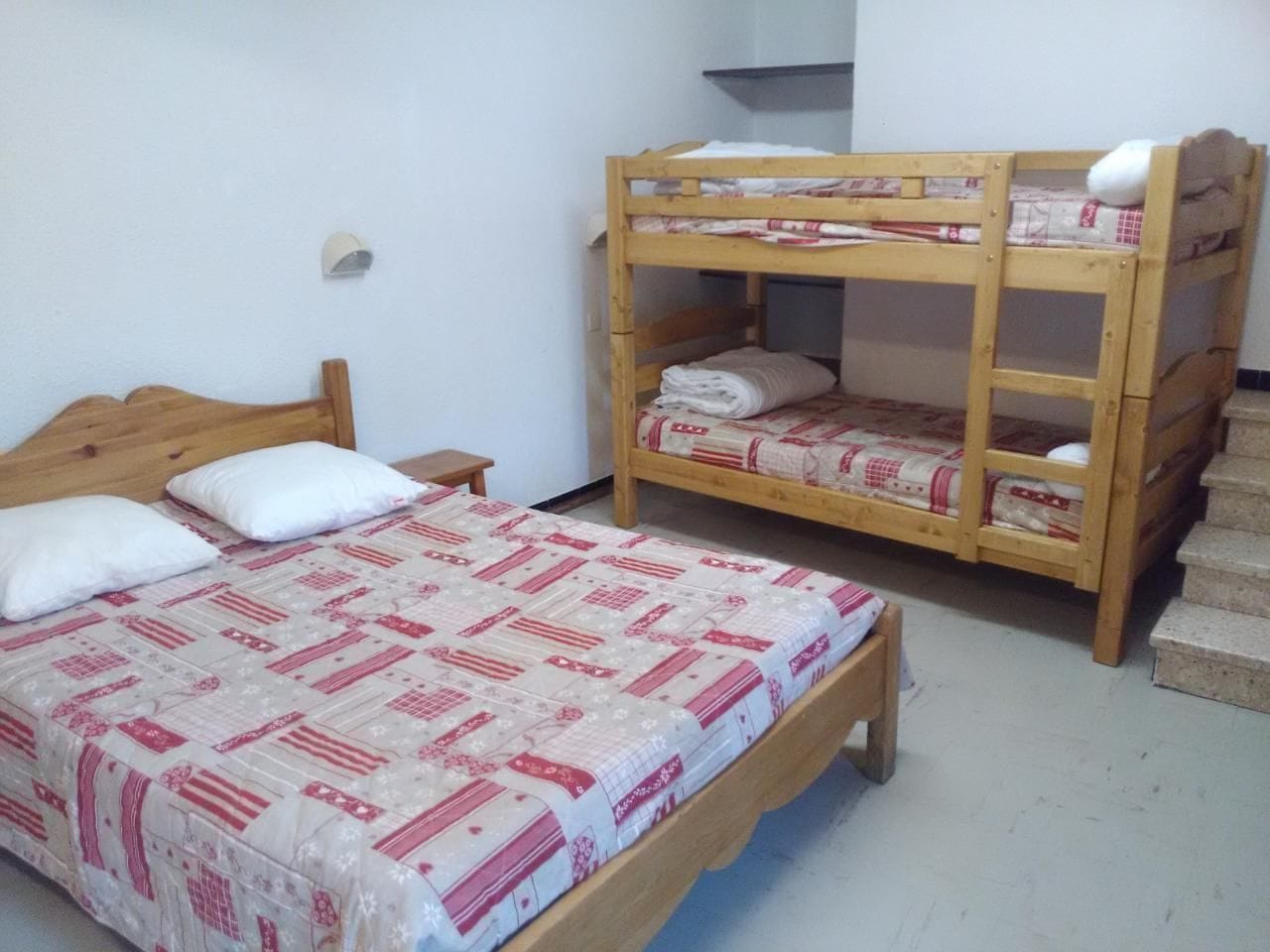 Een kamer met twee houten stapelbedden, waarvan één met een onderbed en één met een bovenbed. Er staat een klein houten nachtkastje met een lamp naast het onderbed.