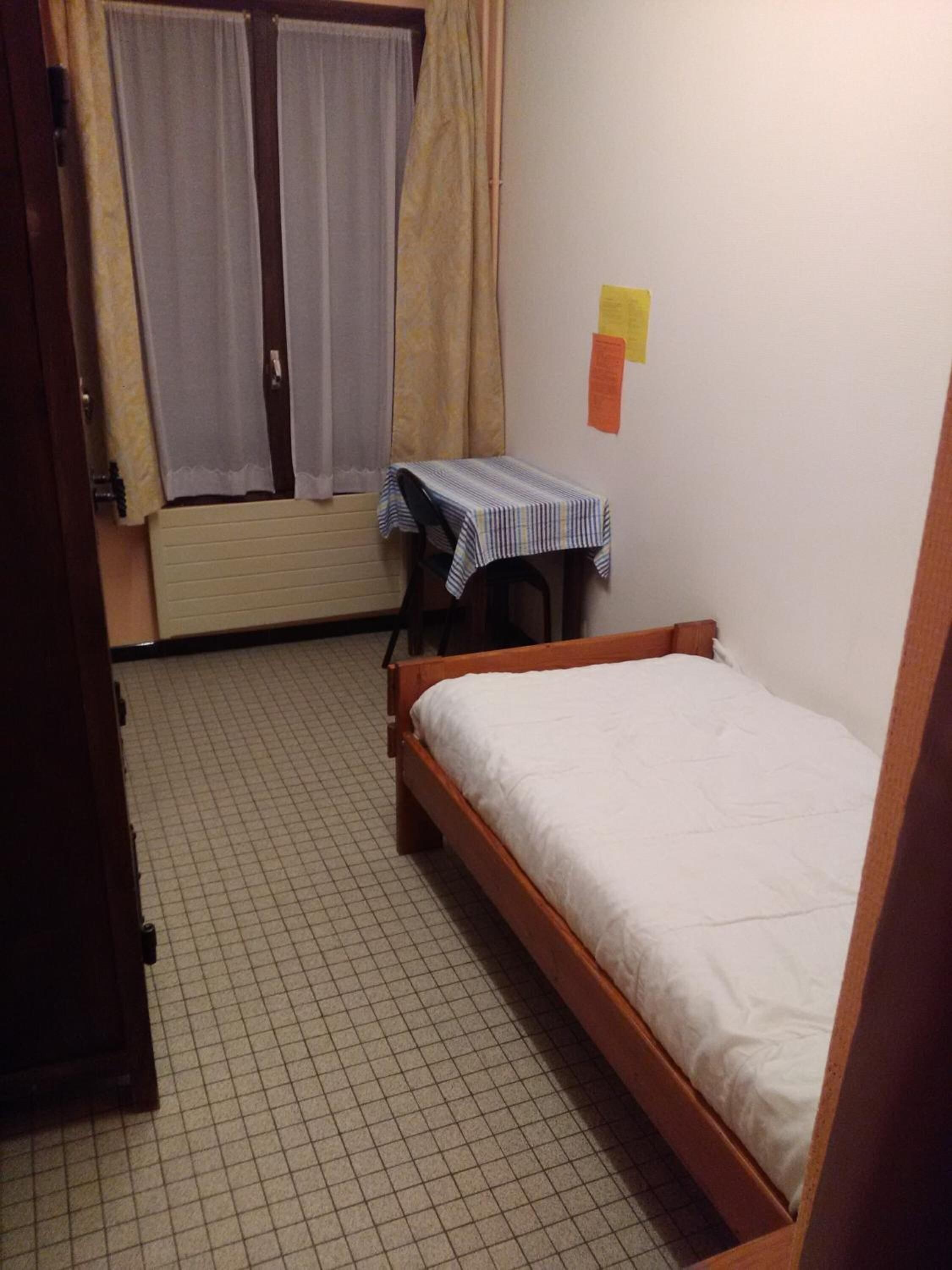 Una habitación pequeña con cama individual, cabecero de madera, una mesita con mantel a rayas, una ventana con cortinas y un radiador.