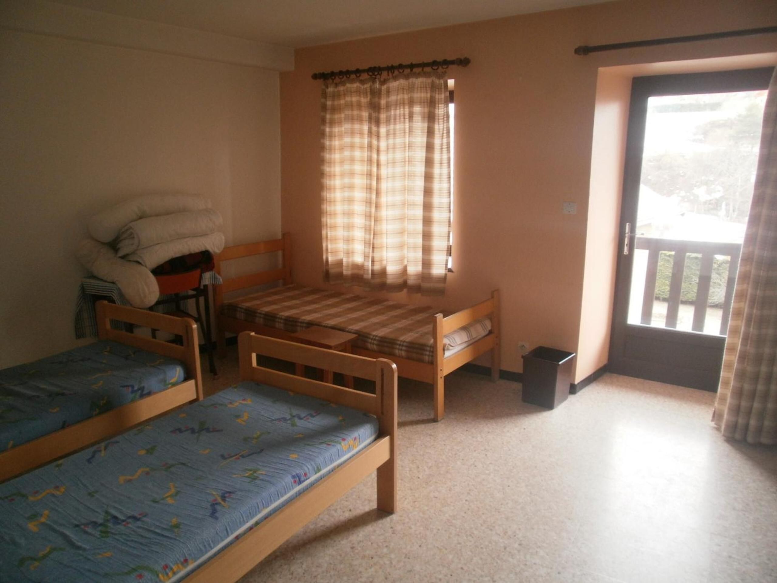 A dormitory room with two single beds, a chair, a small table, a window with curtains, and a door leading to an outdoor area.