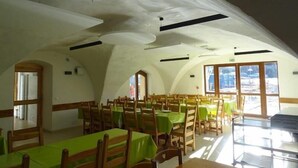 A dining area with wooden tables and chairs, large windows, and a high, arched ceiling. - CapVerb (Guillestre)
