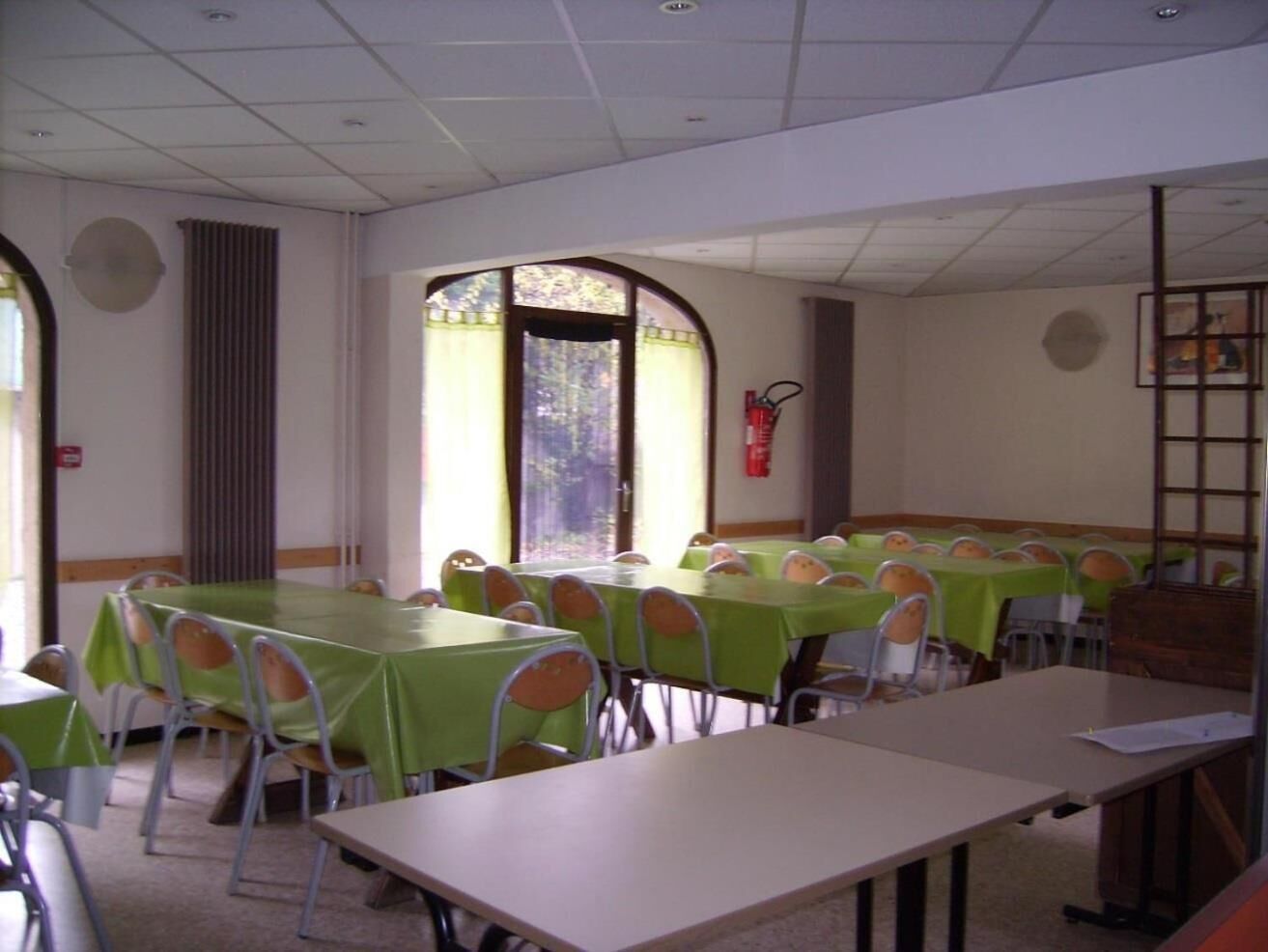 Un salón de conferencias con varias mesas y sillas, ventanas amplias con marcos decorativos y un extintor en la pared.
