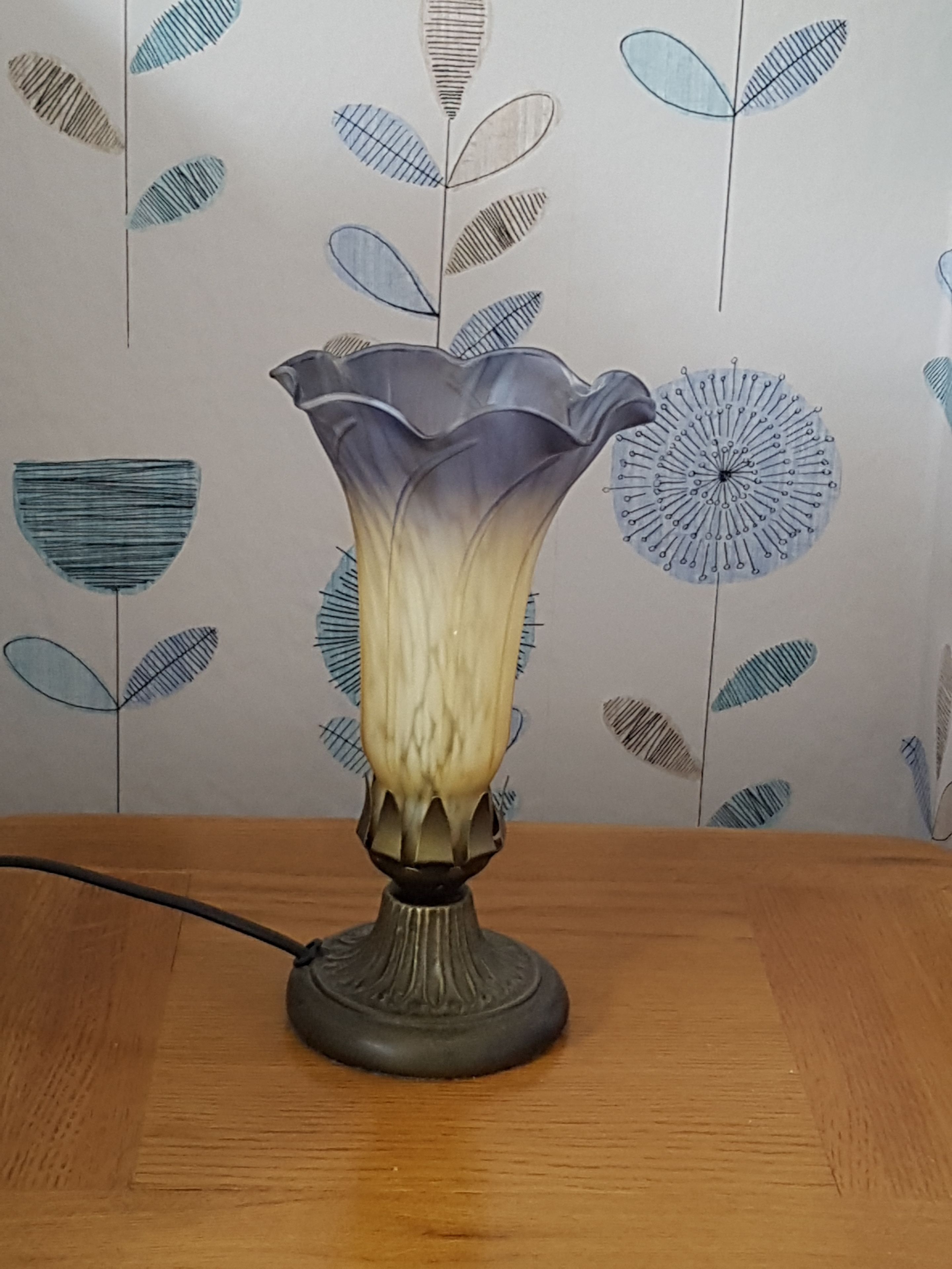 Una lámpara de mesa con base decorativa y pantalla con forma de flor, sobre una mesa de madera frente a un papel tapiz con motivos de hojas.