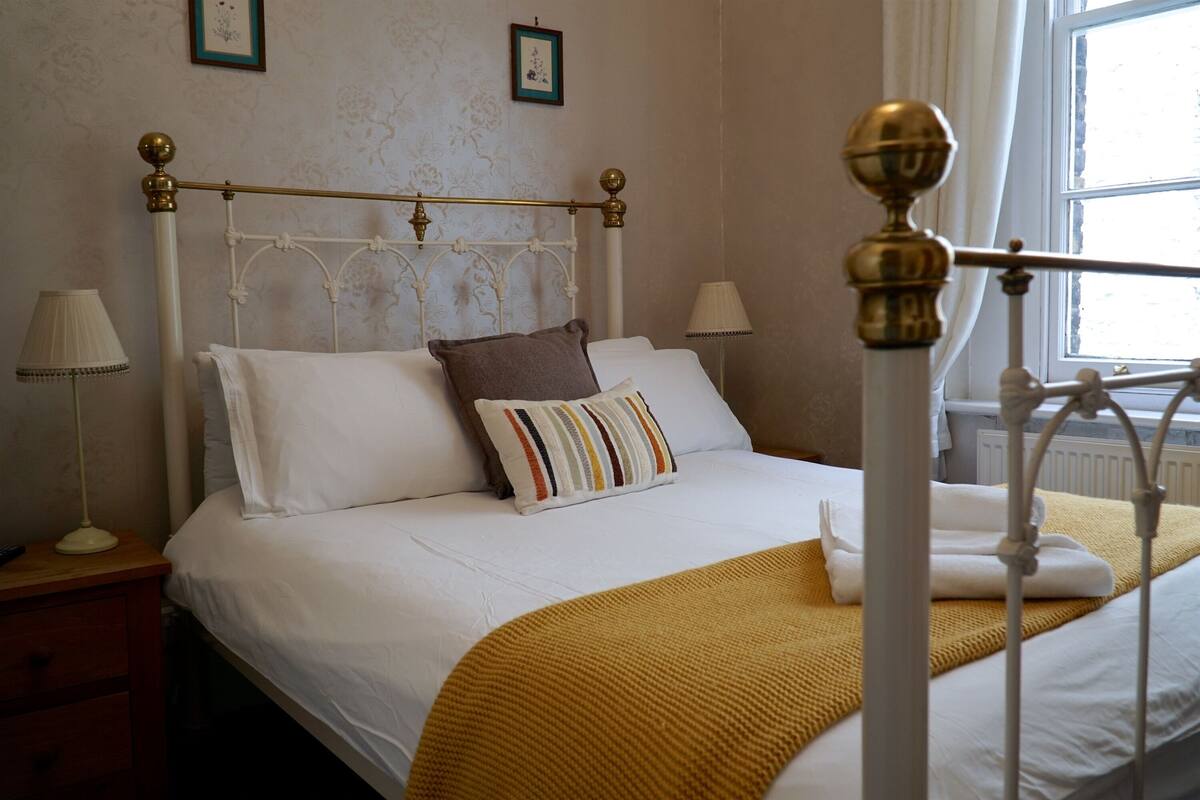 A neatly made bed with white linens, a brown pillow, and a striped pillow. There are two bedside lamps, a wooden nightstand, and a window with curtains.