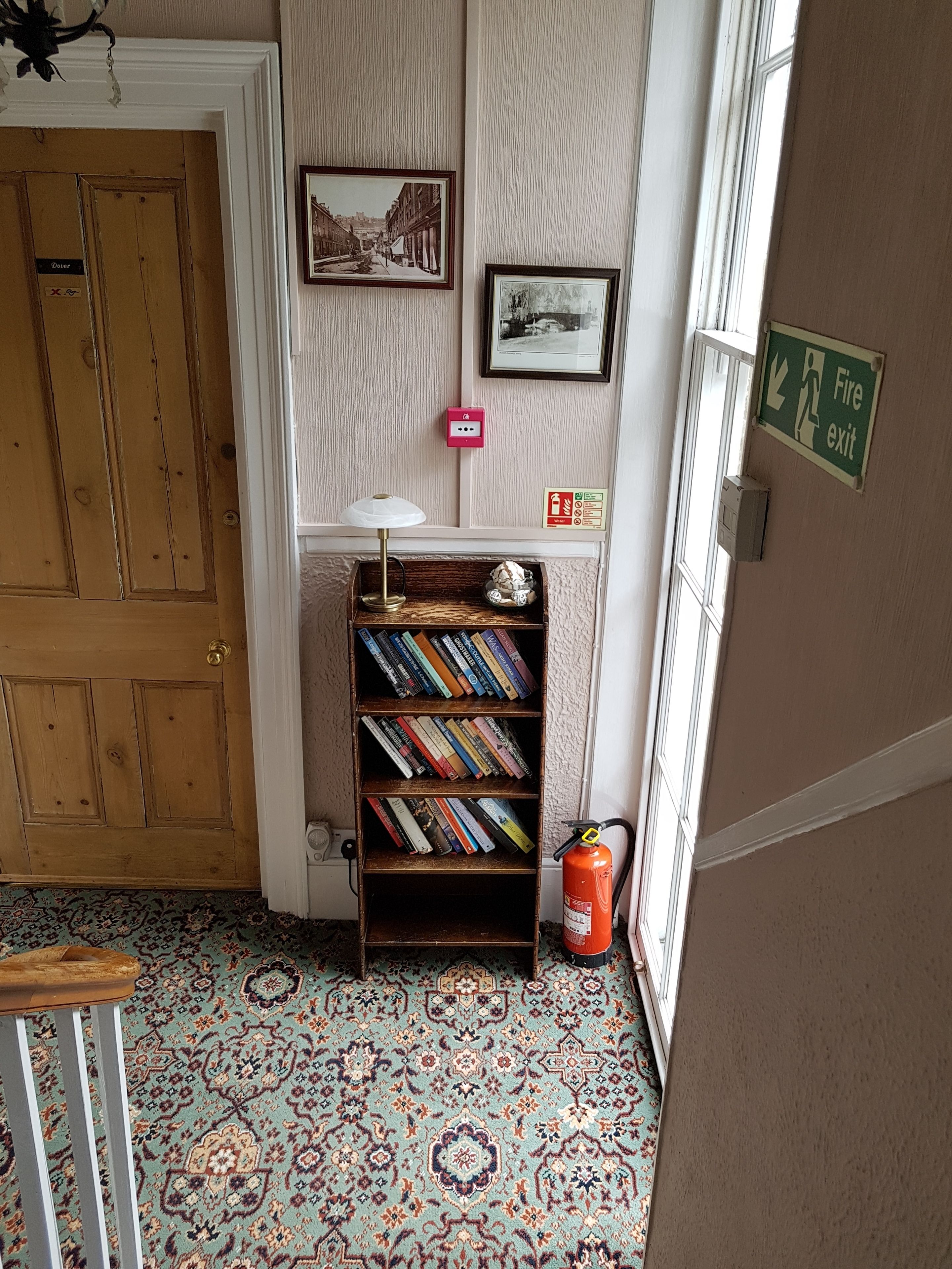 A hallway with a wooden door, a bookshelf with books, a fire exit sign, and patterned carpet.