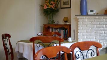 A dining area with wooden chairs and a table covered with a white tablecloth. A bookshelf with books and a vase with flowers are visible in the background.