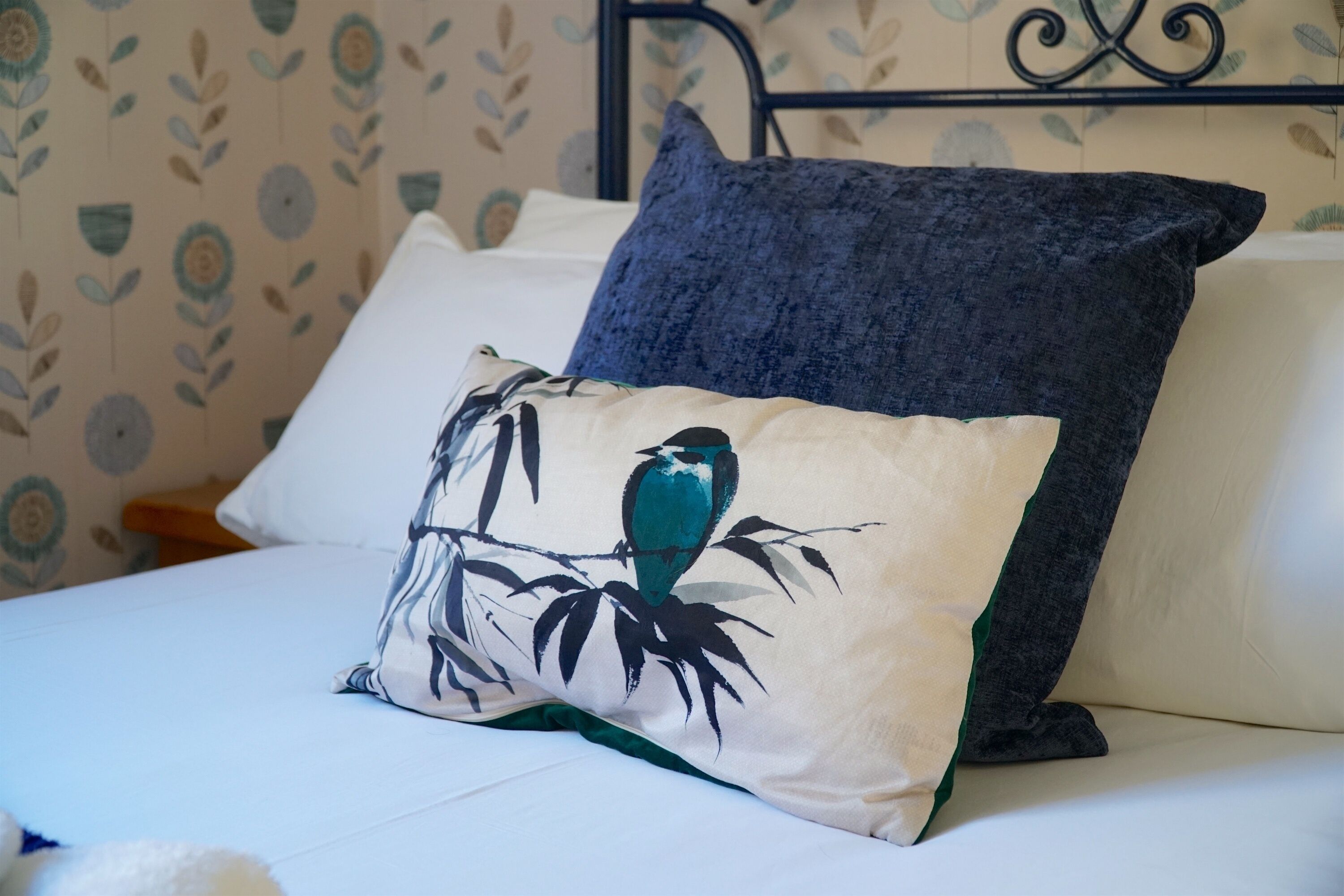 Una cama con ropa de cama blanca, una almohada azul oscuro y una almohada decorativa con un diseño de pájaros y follaje.