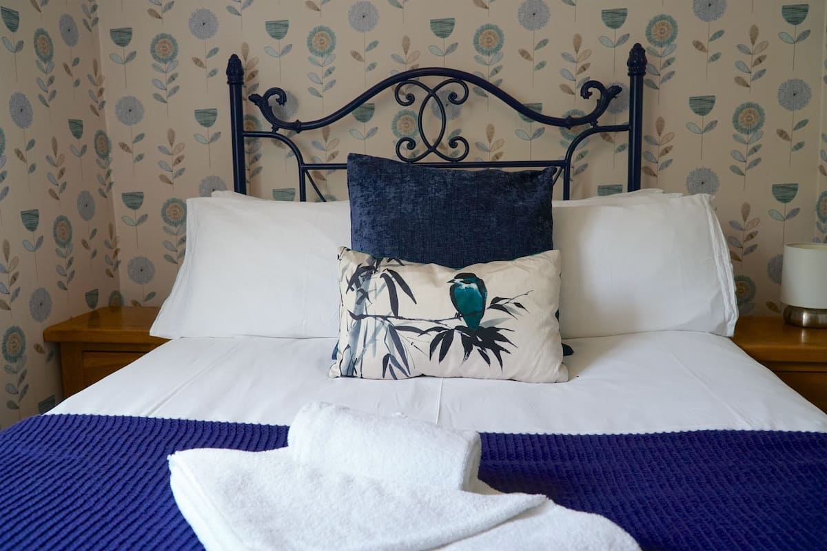 A neatly made bed with a blue blanket, white pillows, and a decorative pillow with a bird design.