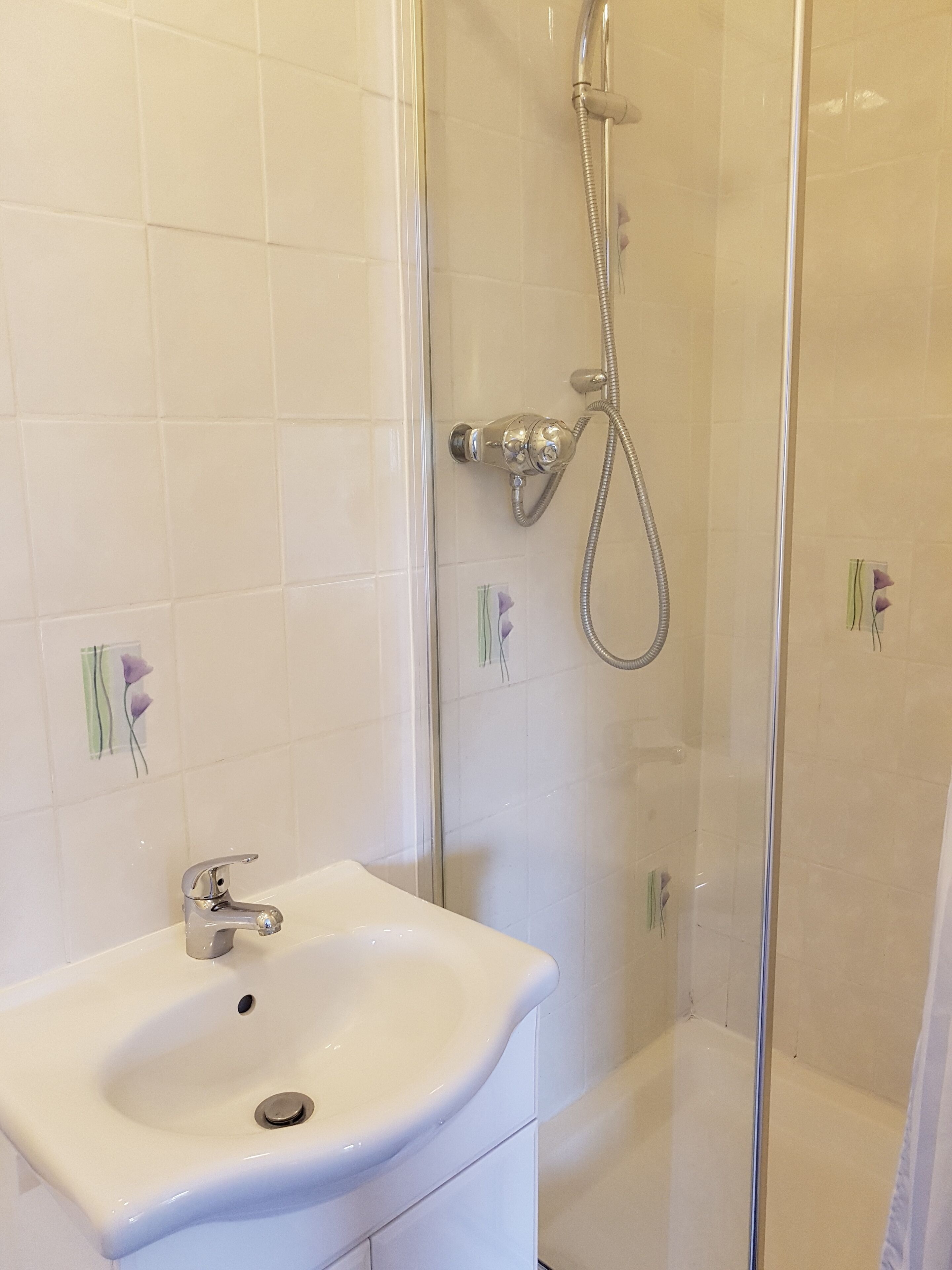 Une salle de bain avec un lavabo blanc, une douche en verre et des carreaux décoratifs ornés de fleurs violettes.