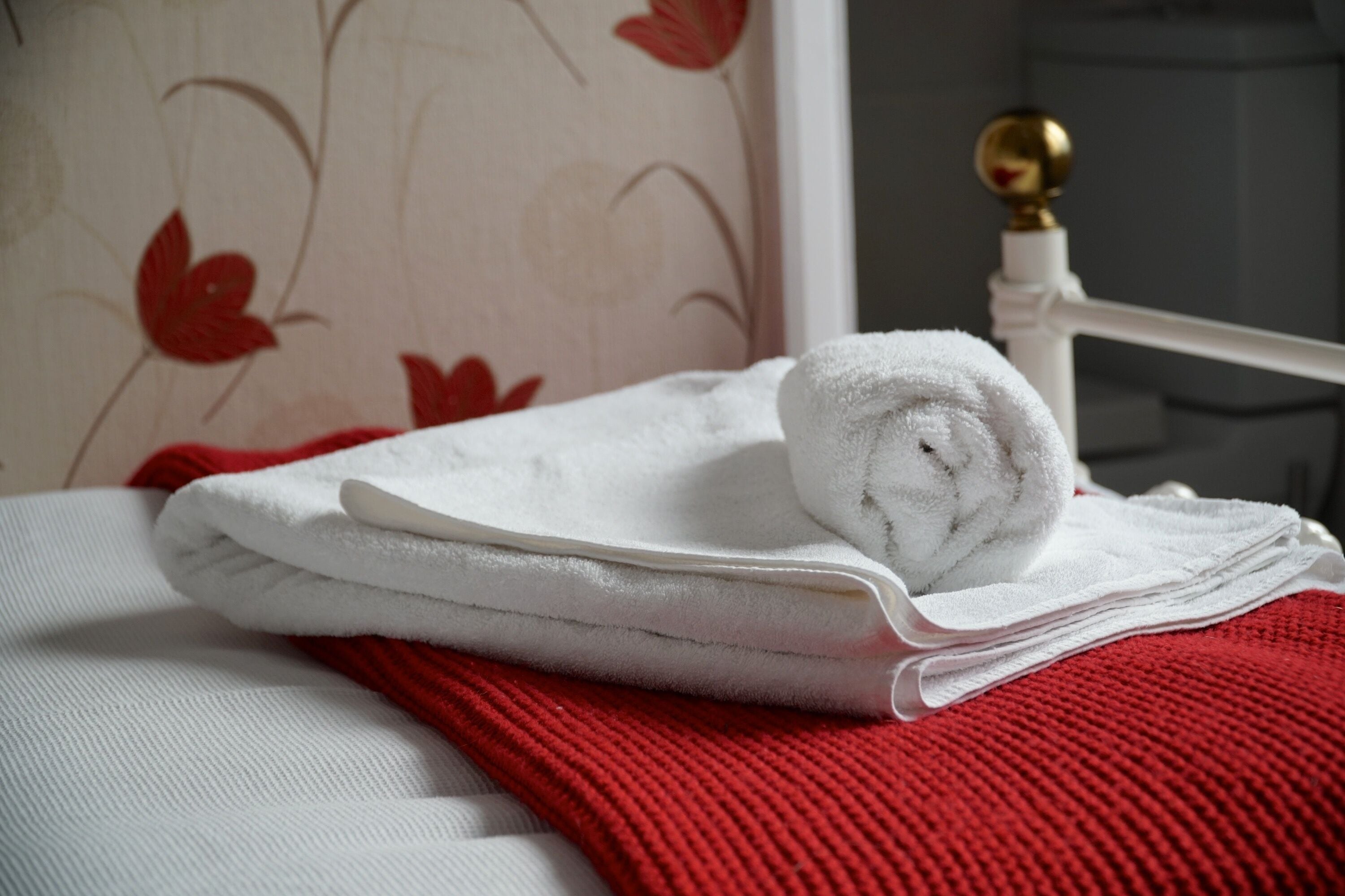 A neatly made bed with white and red linens, a rolled towel, and a gold spherical object on a white chair.