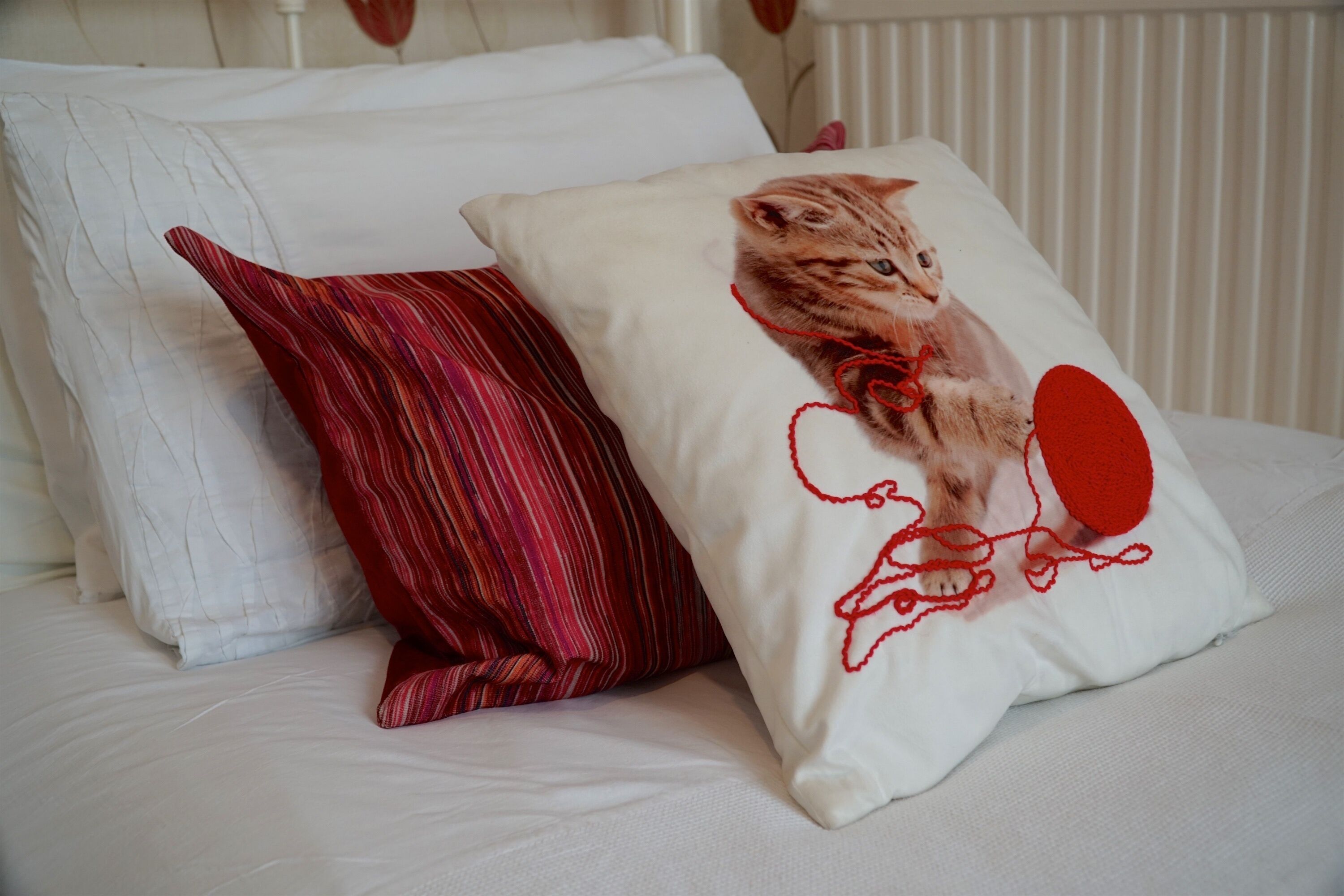 Una cama con ropa de cama blanca y dos almohadas, una con estampado de rayas rojas y blancas y otra con diseño de gato.