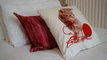 Una cama con ropa de cama blanca y dos almohadas, una con estampado de rayas rojas y blancas y otra con diseño de gato.