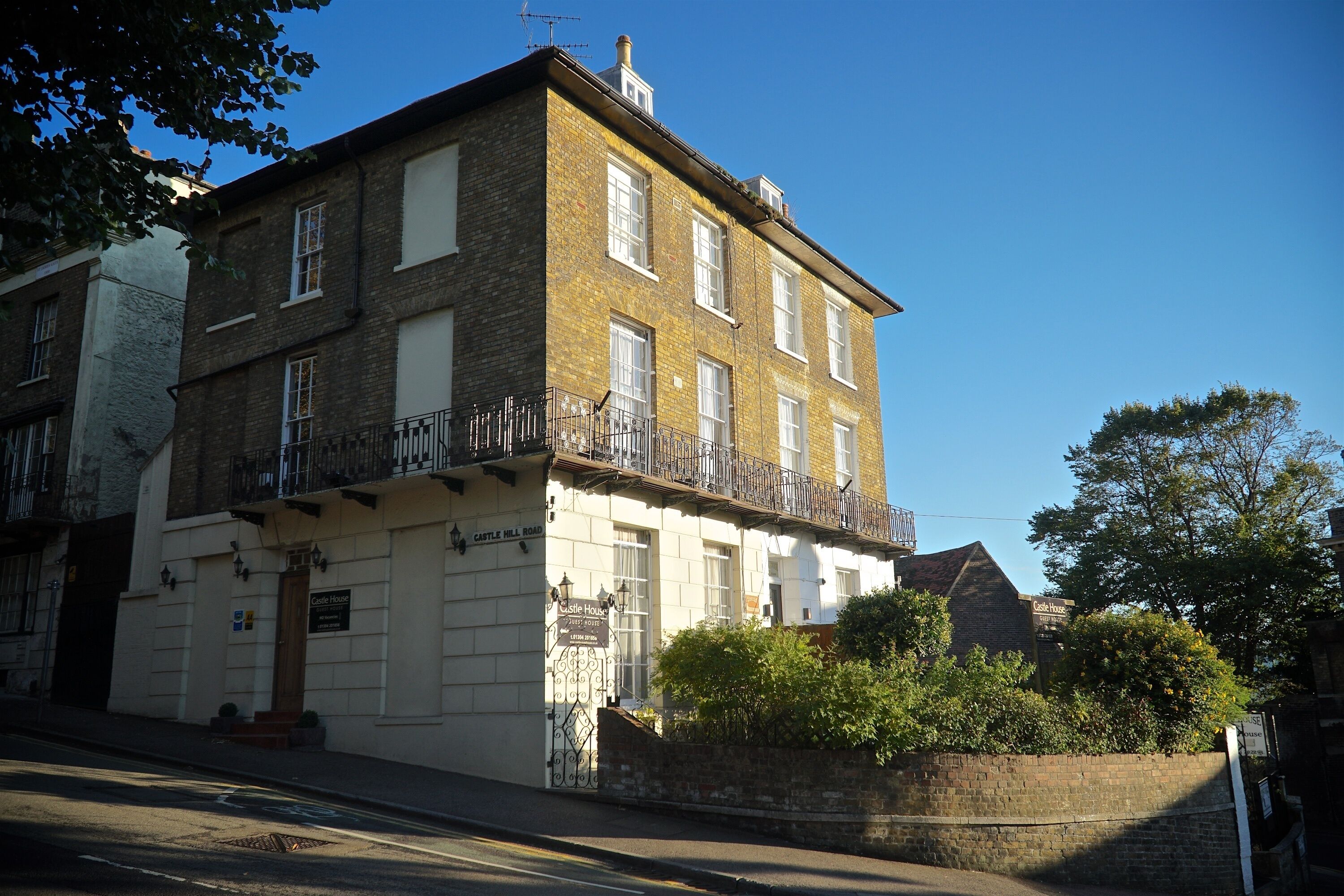 A two-story building with a balcony on the second floor, a sign reading "Castle House," and a brick wall with greenery in front.
