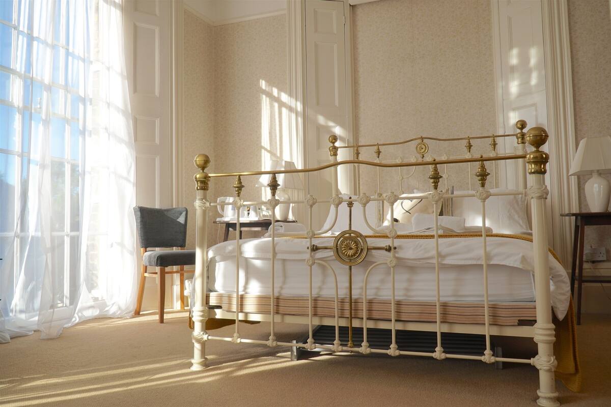 A neatly made bed with a white and gold frame, a chair, and a small table with a lamp.