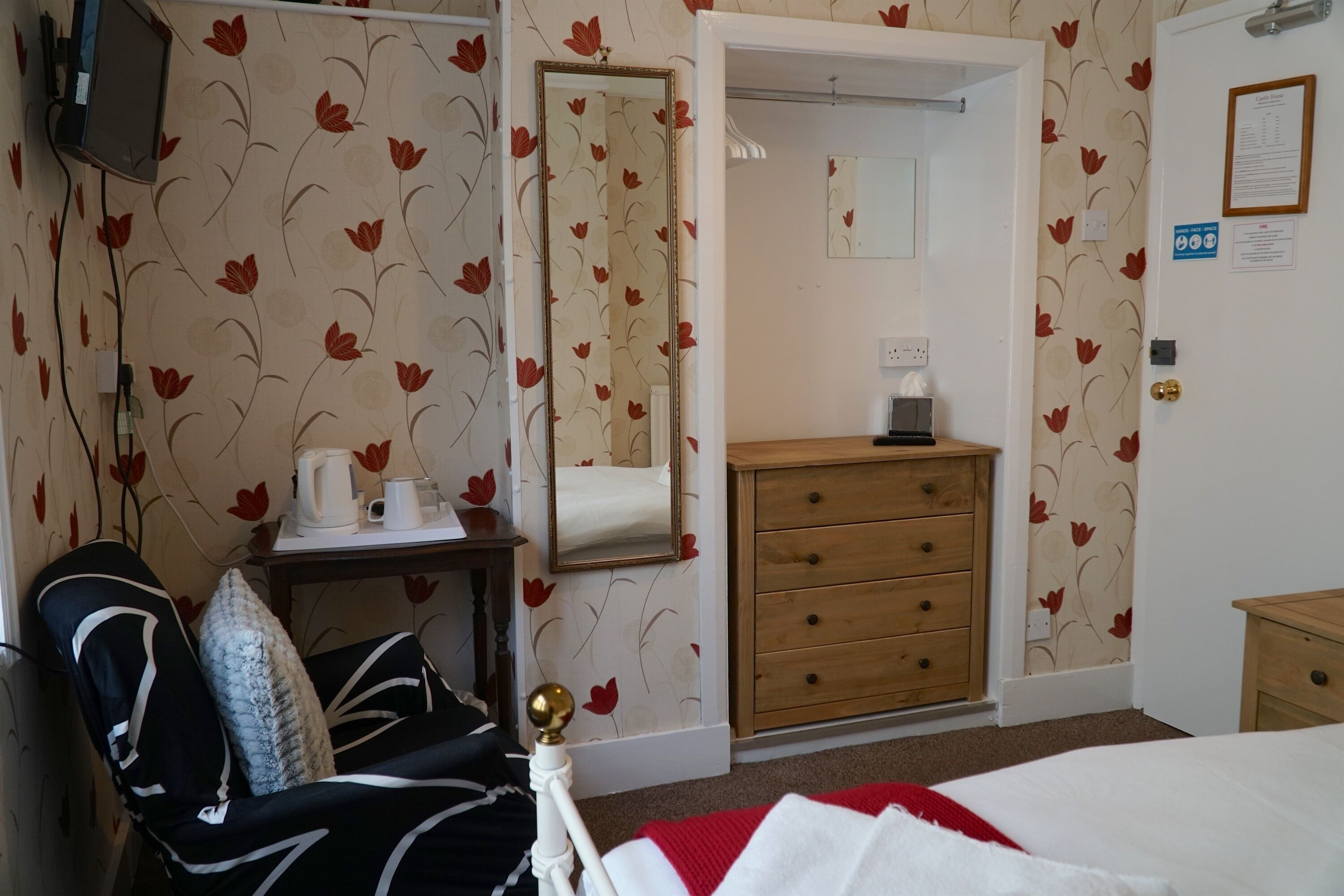 A hotel room with floral wallpaper, a bed with a red blanket, a wooden dresser, a bedside table with a kettle, and a chair with a patterned cushion.