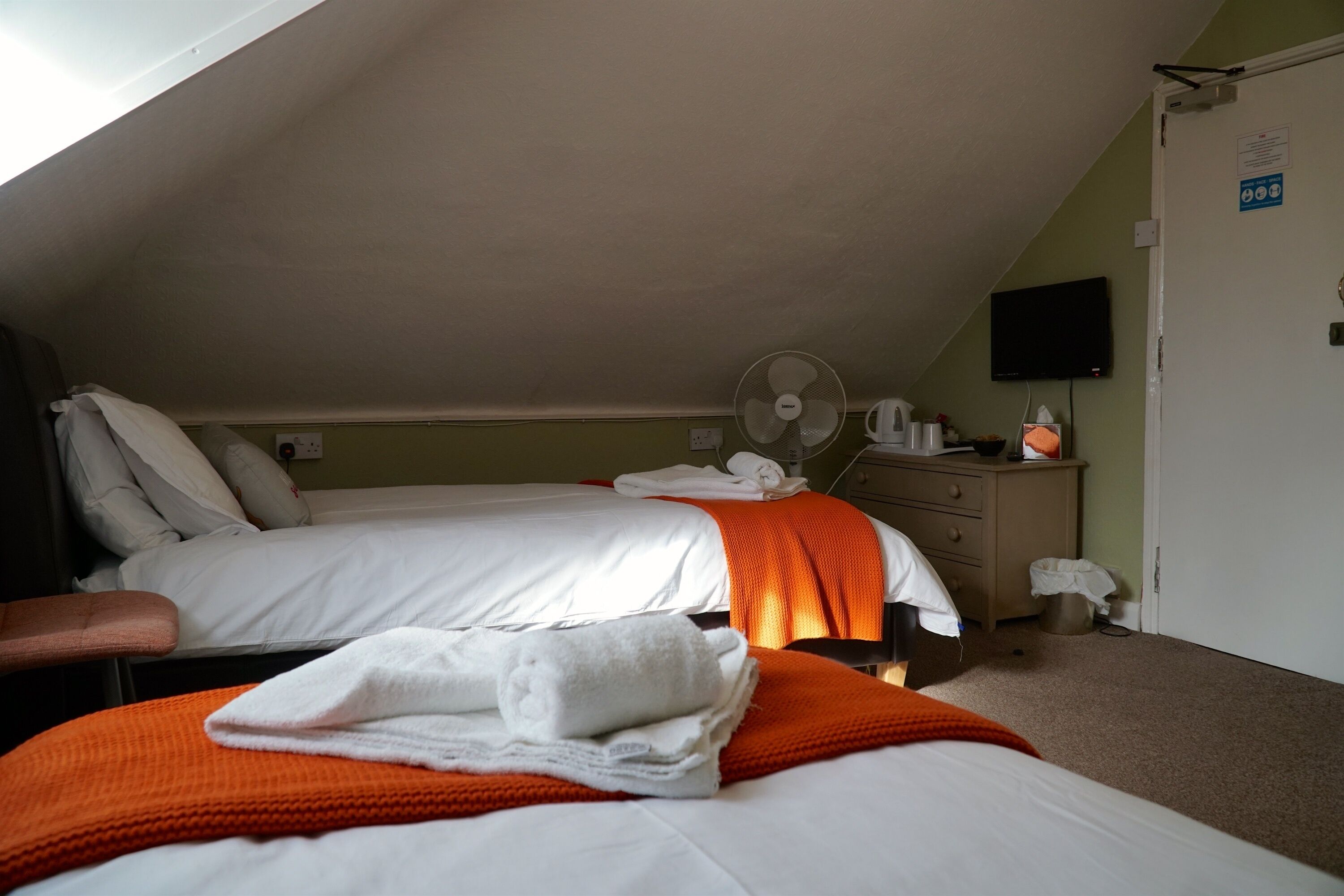 A hotel room with two beds, a fan, a TV, and a small desk with a lamp.