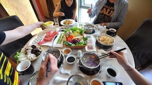 Mesa de comedor con varios platos y una olla caliente, rodeada de gente.