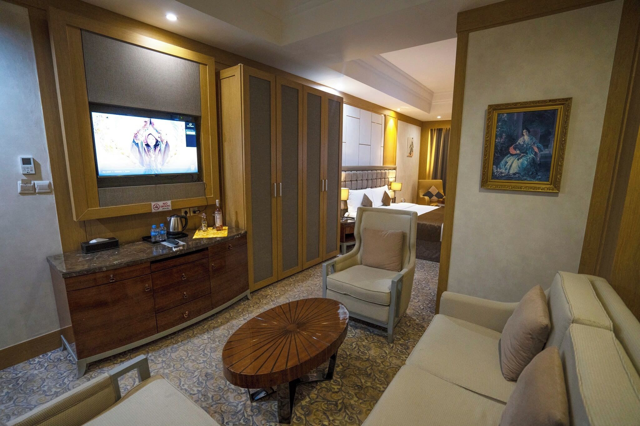 A hotel room with a large bed, a sofa, a dining table, a TV, a wardrobe, and a painting on the wall.