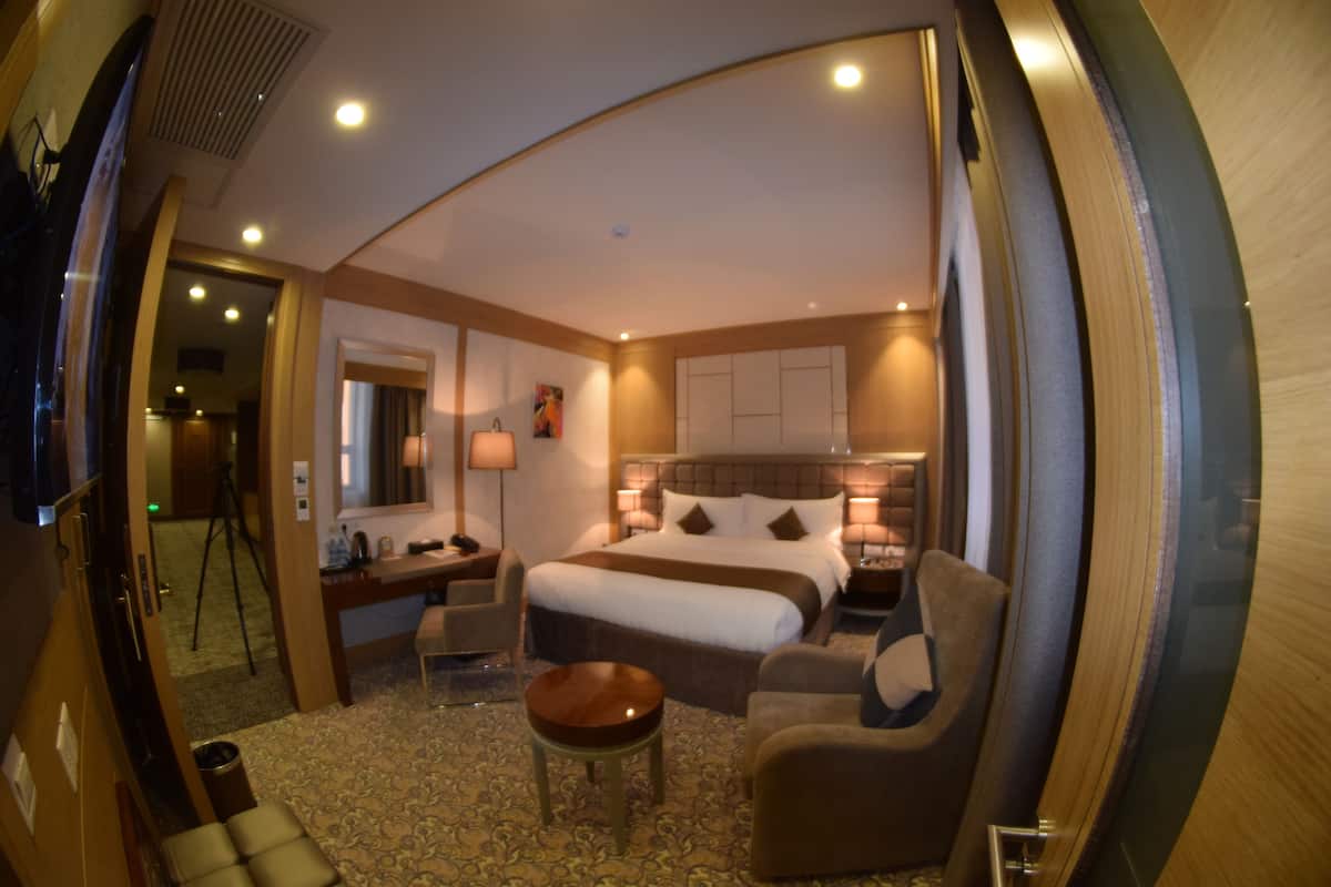 A hotel room with a large bed, a sitting area with a small table, a desk, and a chair. There is a mirror, a lamp, and a window.