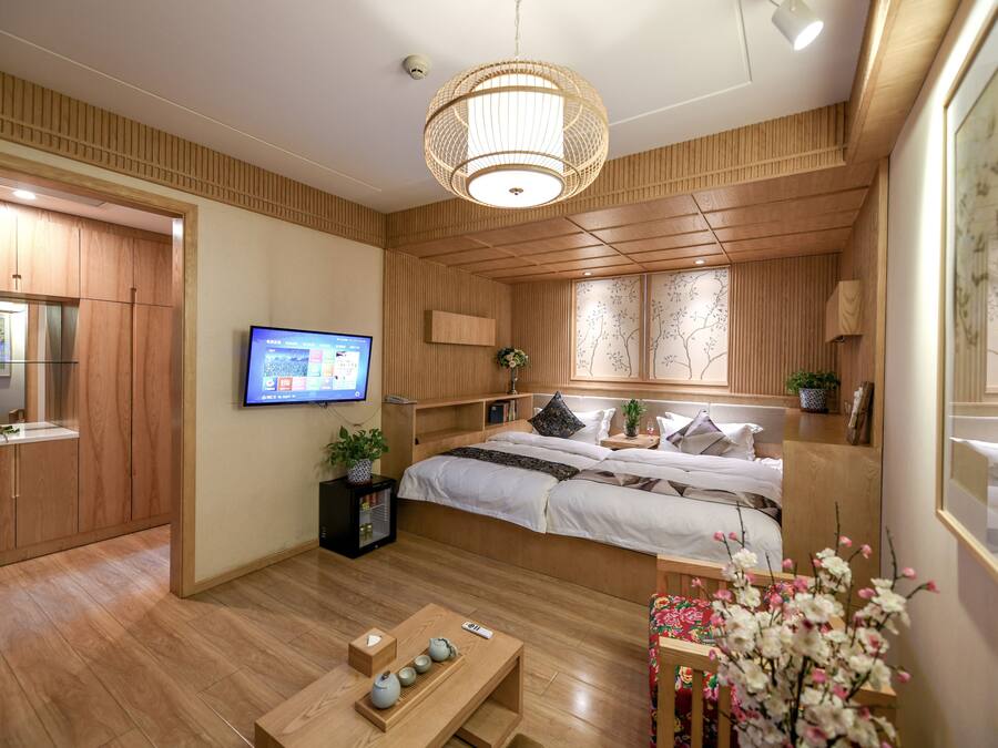 A modern hotel room with a wooden floor, a bed with white bedding, a television, a small desk with a computer, and a large window with a view of trees.