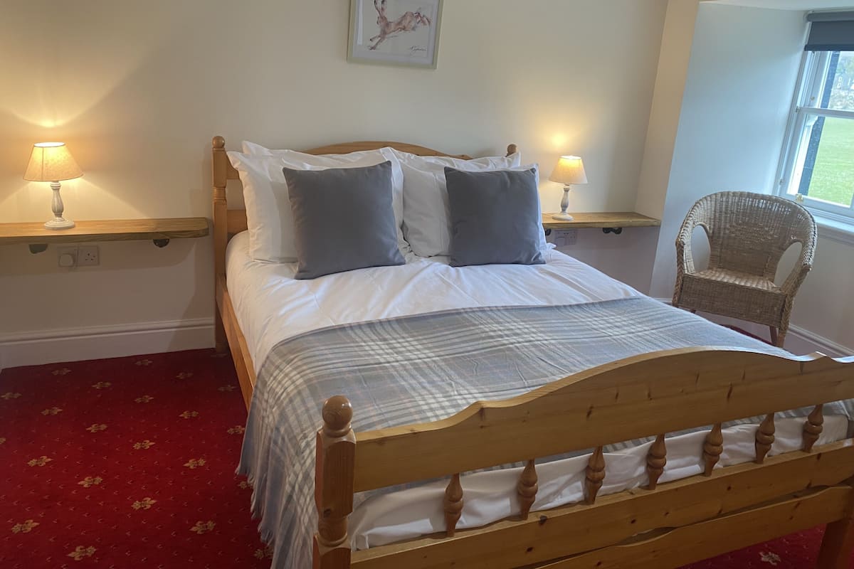 A neatly made bed with a wooden headboard, two bedside tables with lamps, a wicker chair, and a framed picture on the wall.