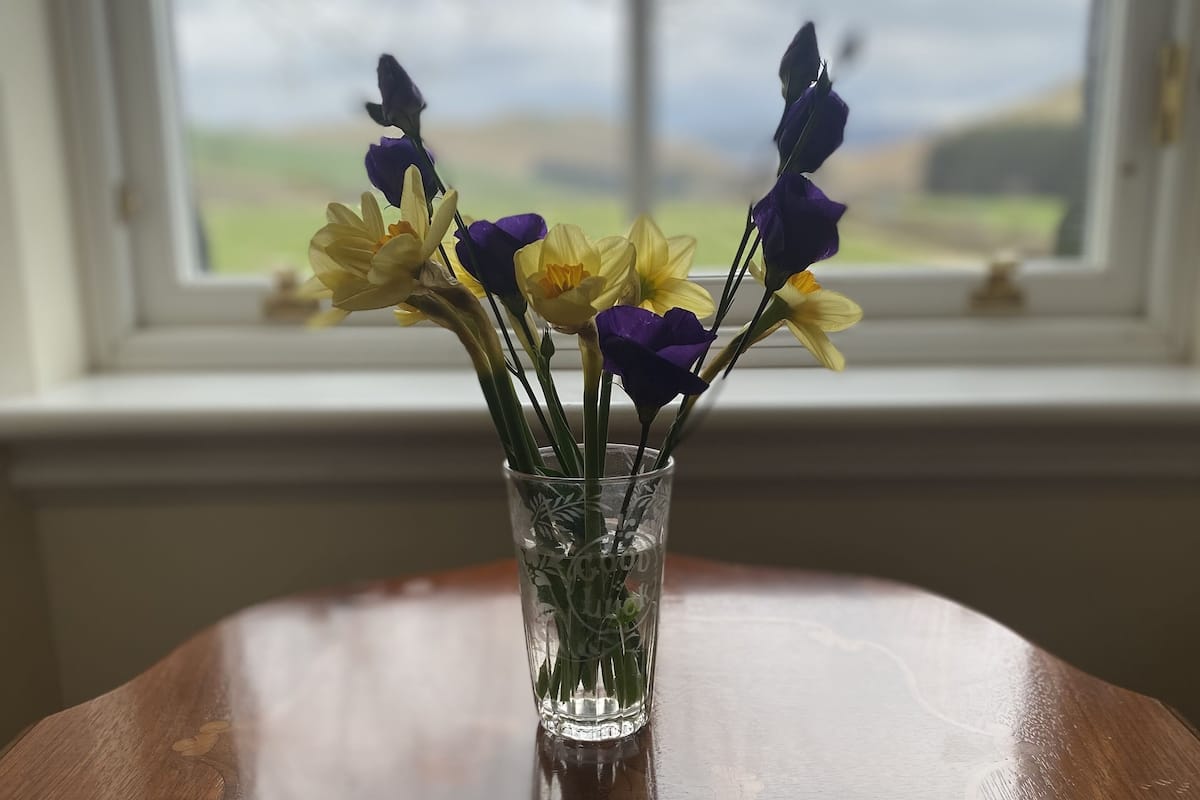 A clear glass vase with yellow and purple flowers on a wooden table by a window.